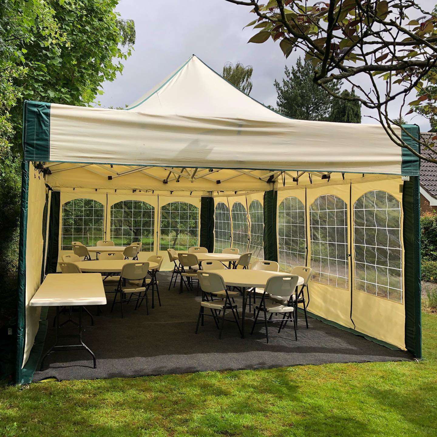 A 4.5m x 4.5m mini marquee gazebo with cream and green side walls, window panels, carpet flooring and round tables with folding chairs set up for a garden party