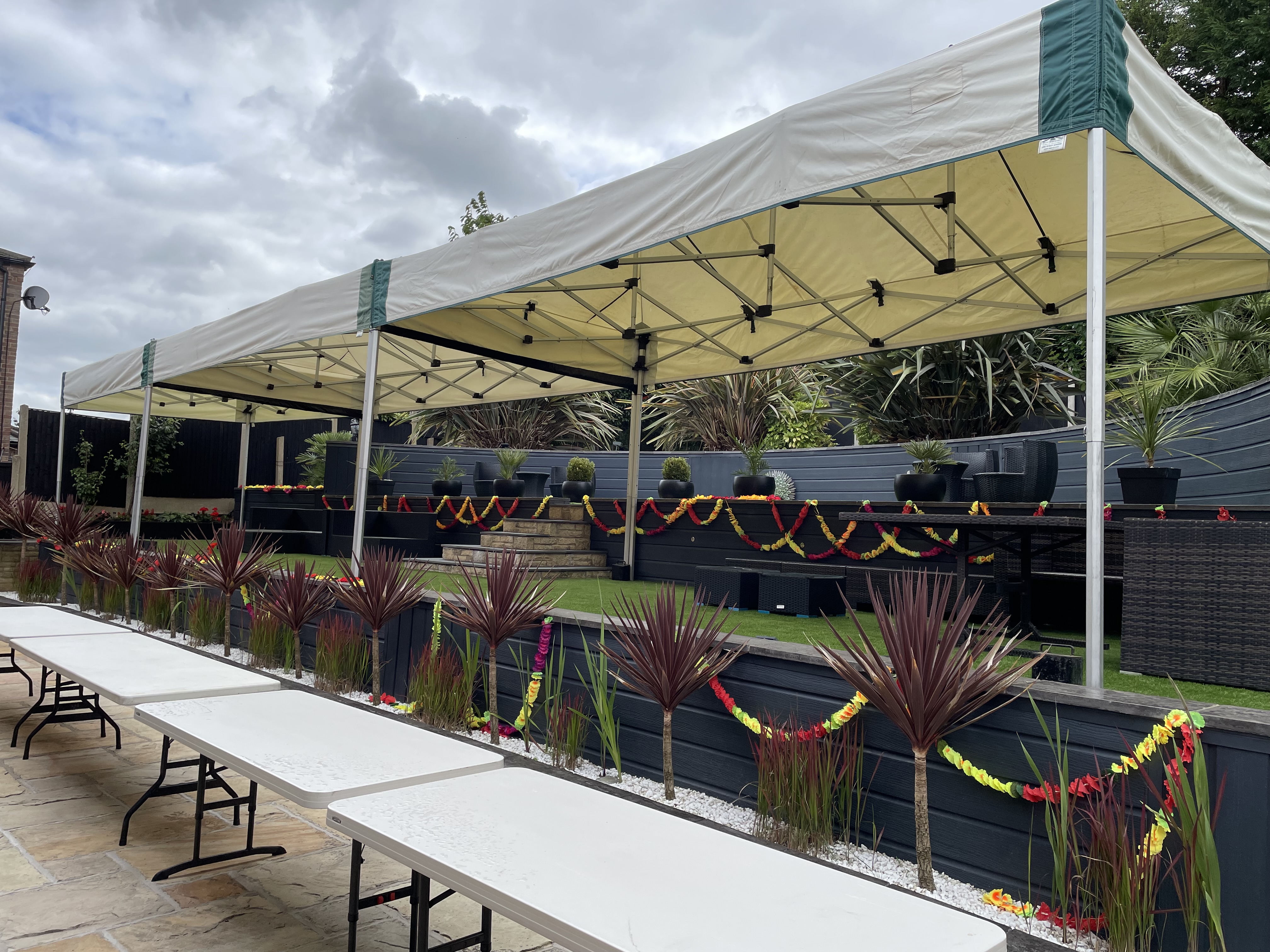 Two linked 6m × 3m mini marquee gazebos at a colourful garden celebration in Ravenshead, decorated with red, yellow and orange floral garlands and long banquet tables