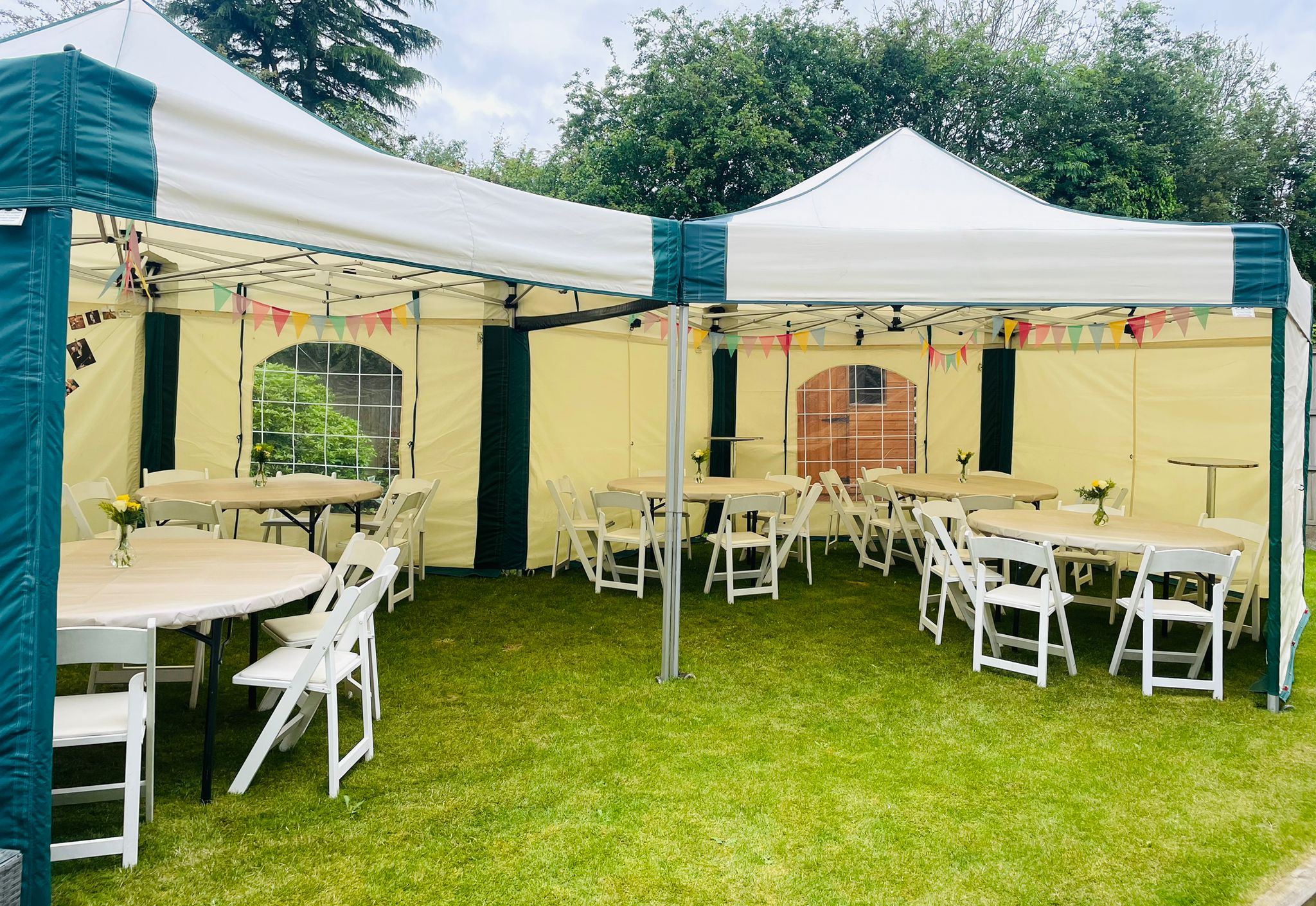 Interior view of two linked hexagonal 6m × 6m mini marquee gazebos beautifully dressed with round tables, white folding chairs, fresh flower centrepieces and colourful bunting