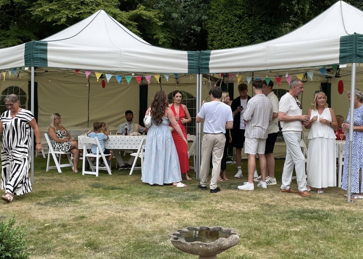 Guests socialising at a summer garden party beneath two linked hexagonal 6m × 6m mini marquee gazebos decorated with colourful bunting, white folding chairs and tables