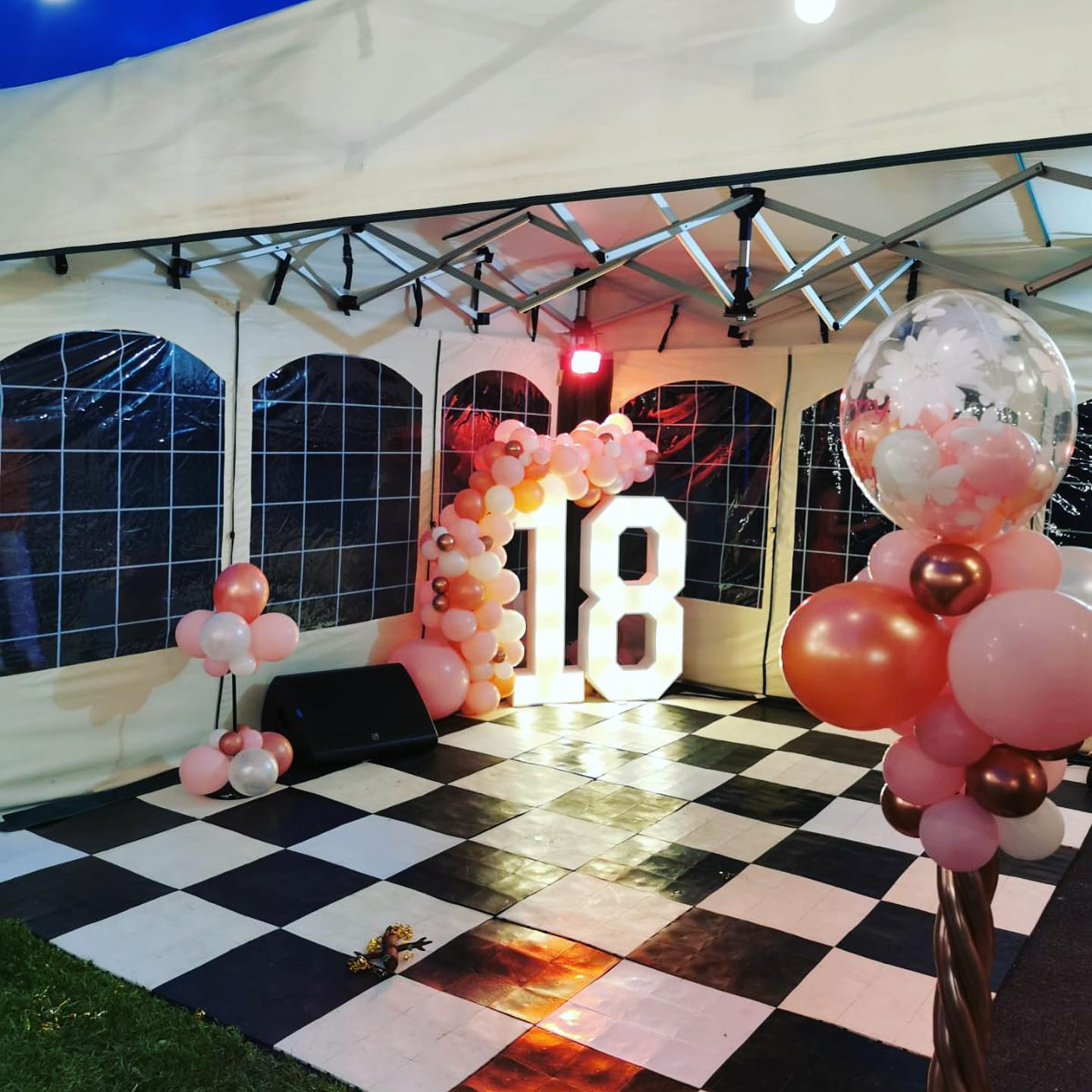 Interior of a 4.5m × 4.5m mini marquee dressed for an 18th birthday party, featuring a black and white chequerboard dance floor, balloon arch, illuminated number 18 and window sidewalls