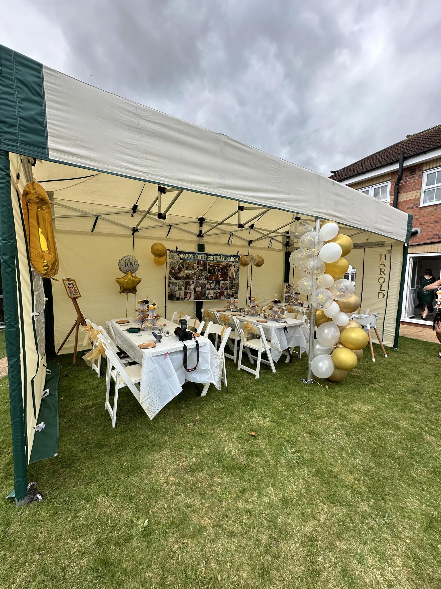 A beautifully decorated 6m x 3m mini marquee gazebo set up for Harold's 100th birthday garden party, with dressed white tables, gold and white balloon arch and personalised decorations