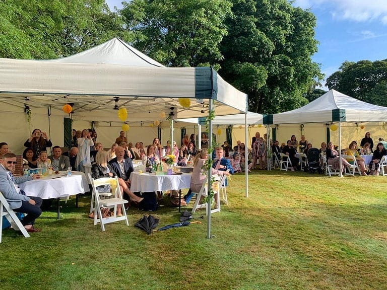 Multiple linked 6m × 6m hexagonal mini marquee gazebos at a large summer garden party, with guests seated at round tables under canopies decorated with colourful bunting and paper lanterns