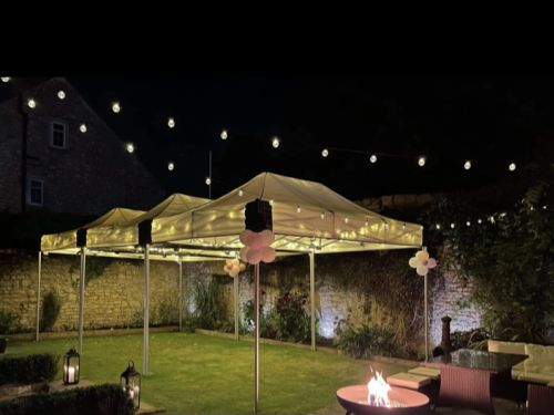 Atmospheric night-time view of two linked mini marquee gazebos forming a 9m × 4.5m covered space, illuminated with festoon lights, pink balloon clusters and a fire pit in a walled garden