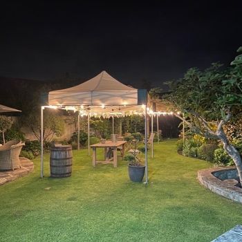 A hexagonal 6m x 6m mini marquee with festoon lights illuminated at night in a private garden, with a rustic barrel and wooden furniture creating an atmospheric evening setting